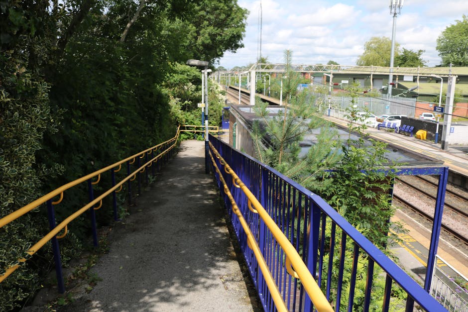 A narrow outdoor pathway with a concrete surface runs alongside a railway station, featuring blue painted metal railings with yellow handrails on both sides. The pathway is partially shaded by overhan