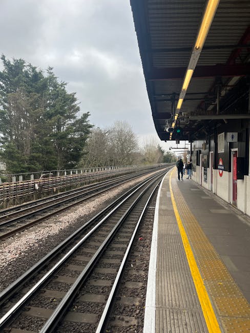A narrow outdoor pathway with a concrete surface runs alongside a railway station, featuring blue painted metal railings with yellow handrails on both sides. The pathway is partially shaded by overhan